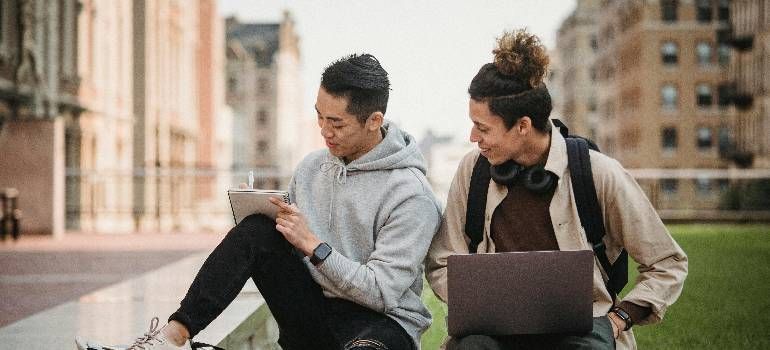 Two people sitting on grass, using a laptop and looking at a phone in an outdoor urban setting