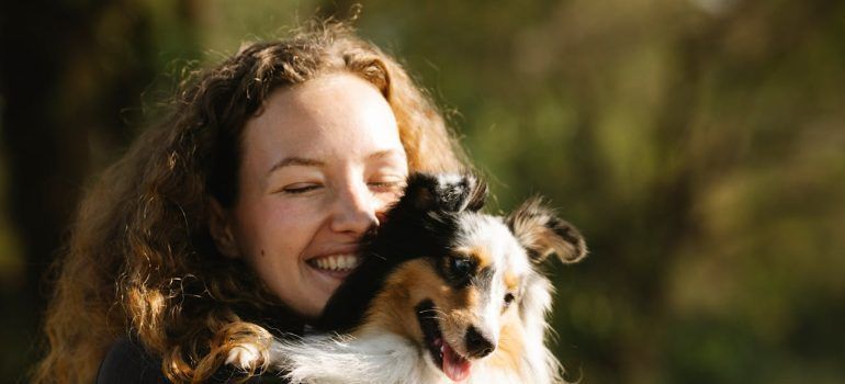 Woman smiling and hugging a black-and-white dog outdoors in warm sunlight