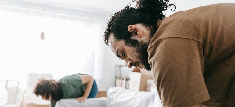 Two people bending over beds in a bright bedroom, one in a brown shirt in the foreground.