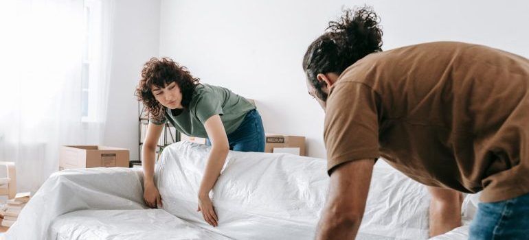 Two people making a bed in a bright bedroom, tucking white sheets over a mattress.