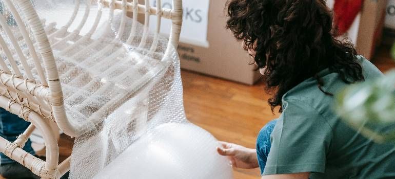 Person crouching beside a white wicker chair indoors, examining a white balloon on a wooden floor.