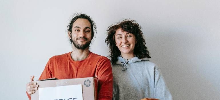 Two smiling people holding a cardboard box against a plain wall