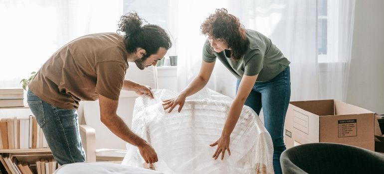 Two people unpacking a white cushion or chair from a box in a bright living room