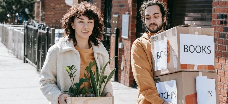 Two people carrying boxes and a potted plant, with a box labeled “BOOKS” in a sunny street setting