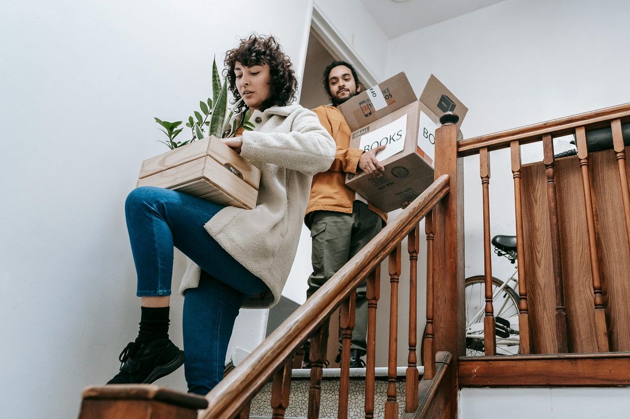 Two people carrying moving boxes down a wooden staircase in a bright home