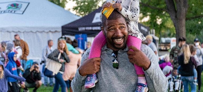 Smiling man carrying a child on his shoulders at an outdoor festival, with tents and people in the background