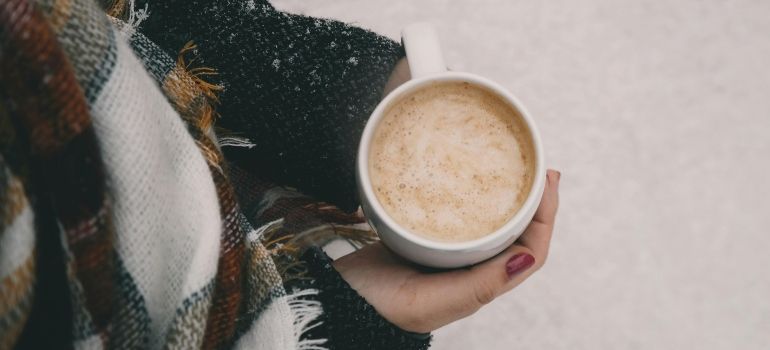 Hand holding a cup of coffee beside a plaid scarf, seen from above on a light surface