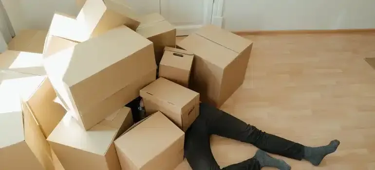 Person buried under many cardboard boxes in a room, legs sticking out on a wooden floor