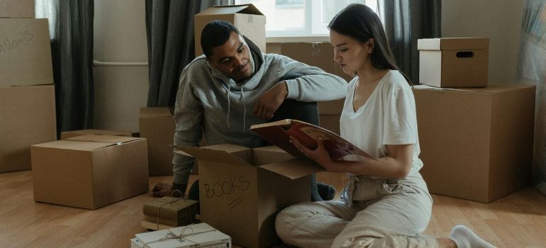Two people unpacking books on the floor of a moving apartment, surrounded by cardboard boxes