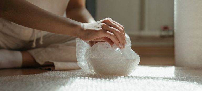 Hands pressing bubble wrap on a sunlit floor beside a seated person in soft clothing