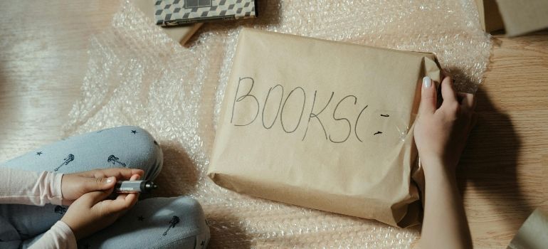 Brown paper bag labeled “BOOKS” on a bed with a blue stuffed animal nearby, viewed from above
