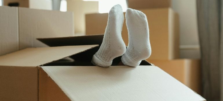 Person's socked feet sticking out of an open cardboard box amid moving boxes