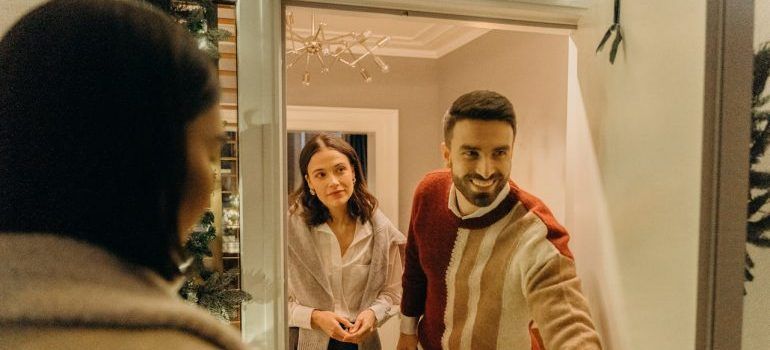Two people smiling at the front door, one opening it from inside a warmly lit home