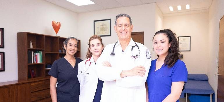 Medical staff posing in a clinic hallway, including a doctor in a white coat and two nurses in blue uniforms.
