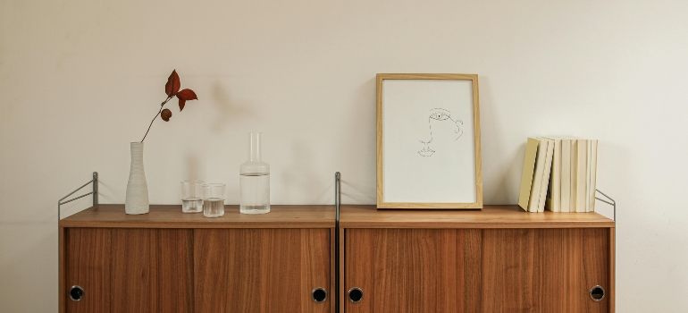 Minimal wooden cabinet with a vase, framed art, glass, and books against a plain wall