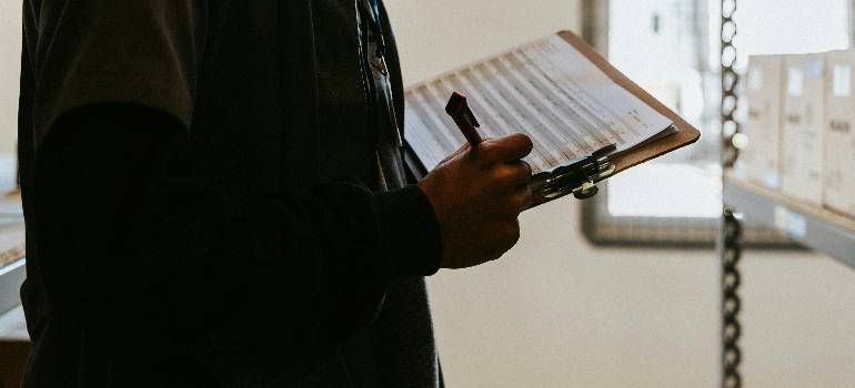 Person holding a clipboard and pen near a window, reviewing notes in a dimly lit room