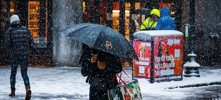 Snowy city street with pedestrians, one holding a black umbrella, beside red postal boxes and lit storefronts