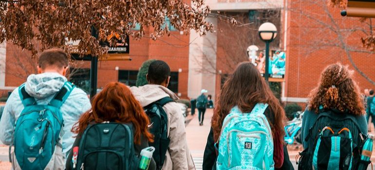 Students with backpacks walking along a tree-lined campus path between brick buildings
