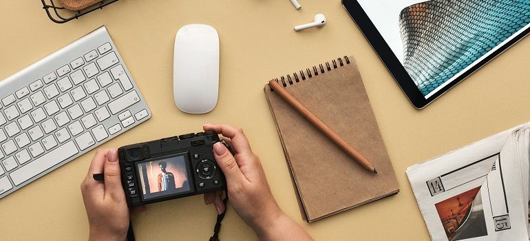 Hands holding a camera on a desk with keyboard, mouse, notebook, pencil, and magazine.