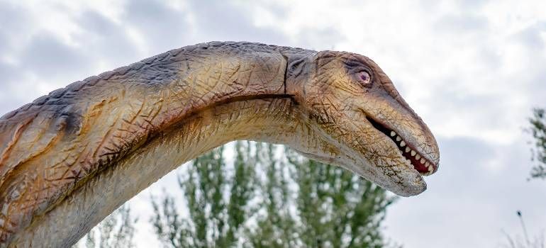 Close-up of a long-necked dinosaur statue against a cloudy sky with trees in the background