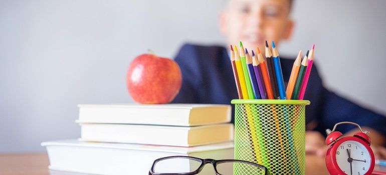 School desk with stacked books, apple, pencils in a cup, glasses, and an alarm clock