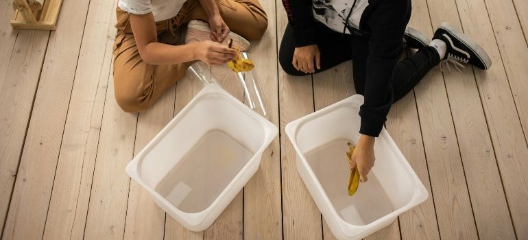 Two people on a wooden floor place liquid into two white plastic bins