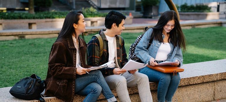 Three people sit on a bench outdoors, studying papers in a grassy campus area.
