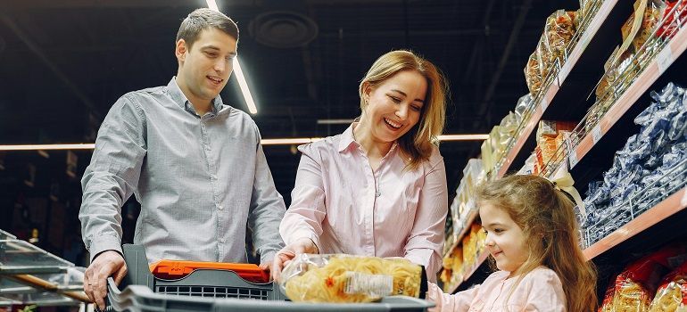 Family shopping in a grocery store, smiling by a cart loaded with produce and bread.