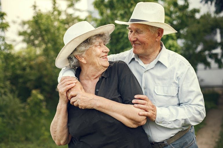Two older adults in white hats smiling and embracing outdoors in a green field