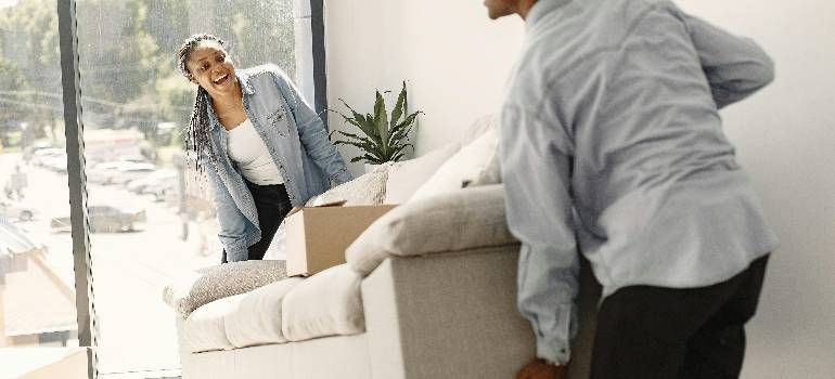 Two people moving a beige couch in a bright living room with large windows