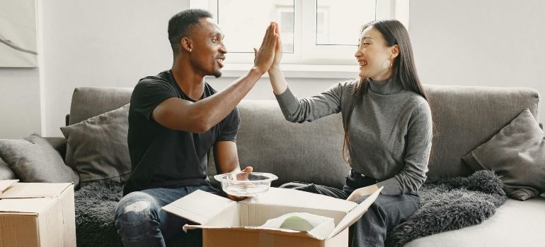 Two people high-five on a couch beside an open takeout box in a bright living room.
