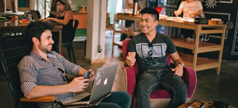 Two coworkers chatting in a café lounge, one using a laptop and the other holding a tablet.