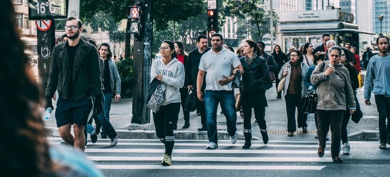 Pedestrians crossing a city street at a crosswalk, with storefronts and trees in the background.