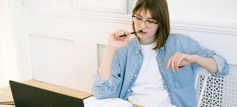 Person in glasses sits at a laptop, thinking with a pen near their mouth in a bright room.