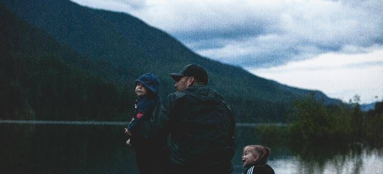 Three people in jackets by a lake at dusk, with mountains and cloudy sky in the background
