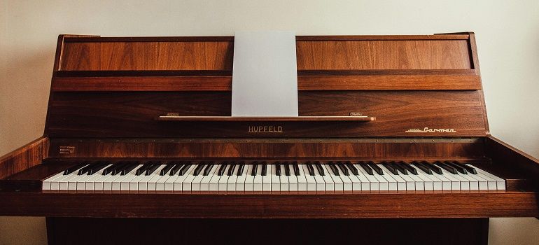 Wooden upright piano with closed music stand and exposed keyboard, against a plain wall