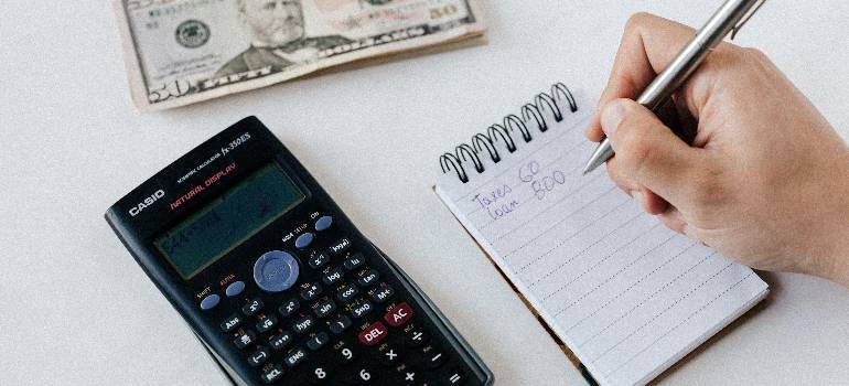 Hand writing in a spiral notebook beside a calculator and cash on a white desk