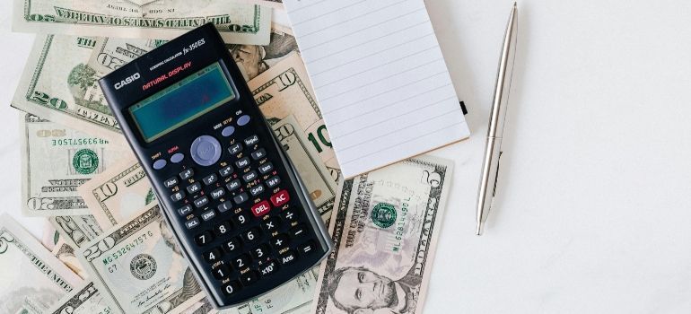 Calculator on scattered dollar bills beside a notepad and pen on a white background