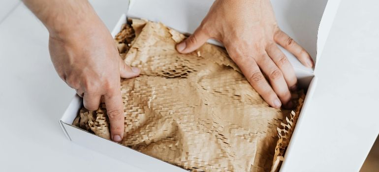 Hands pressing tan packing paper into a white box with brown shredded fill inside