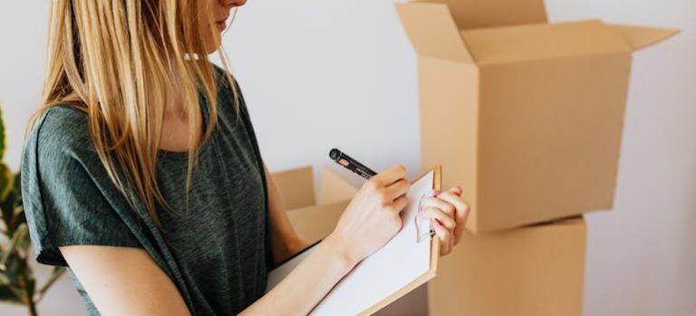 Person writing on a clipboard beside stacked cardboard boxes in a moving or packing scene