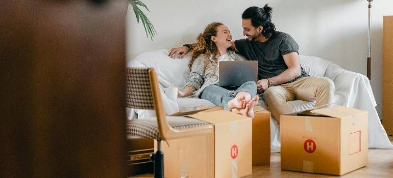 Couple sitting among moving boxes on a couch, smiling at a laptop in a bright living room.