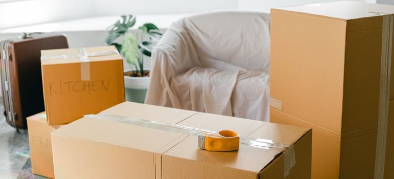 Packed moving boxes in a bright room with a white chair and yellow tape measure on a box