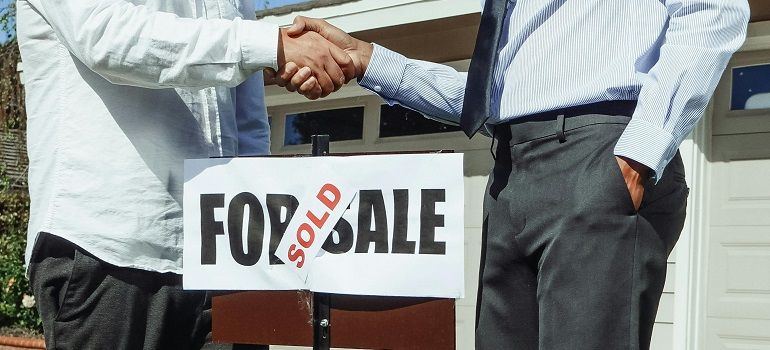 Two people shaking hands beside a house-for-sale sign with a red SOLD banner.