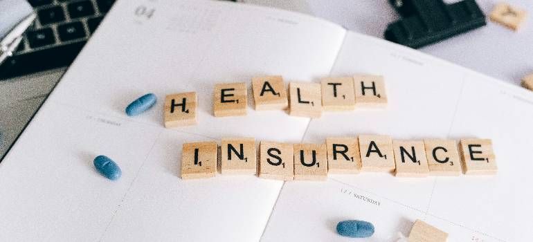 Wooden tiles spelling “HEALTH INSURANCE” with blue pills on a white desk