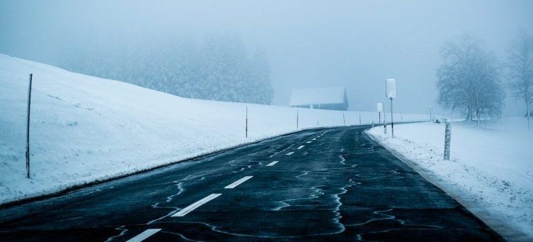 Snow-covered road stretching into a foggy winter landscape with bare trees and low visibility