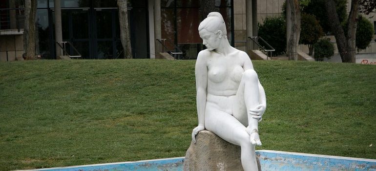 White statue of a seated figure in a fountain, with a grassy lawn and building in the background