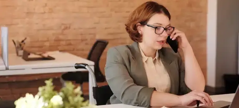 Person in glasses talking on a phone at a desk in an office, working on a laptop.