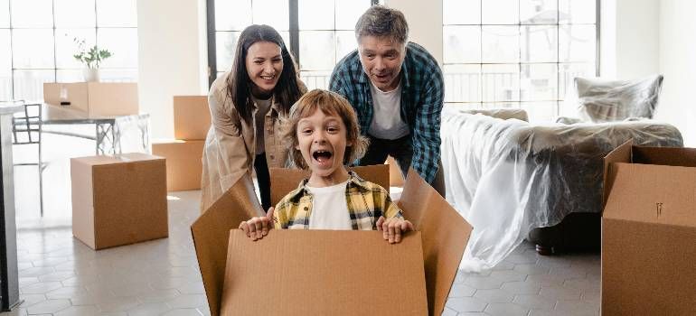 Family smiling in a bright living room, with a child sitting in a cardboard box amid moving boxes.