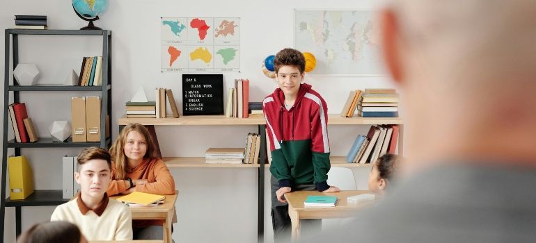 Classroom scene with students listening at desks and a teacher in the foreground with a globe on a shelf.