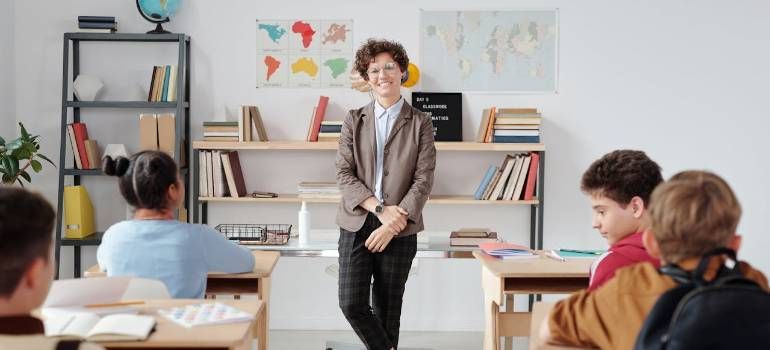 Teacher stands in a classroom as students sit at desks, with bookshelves and maps behind her.
